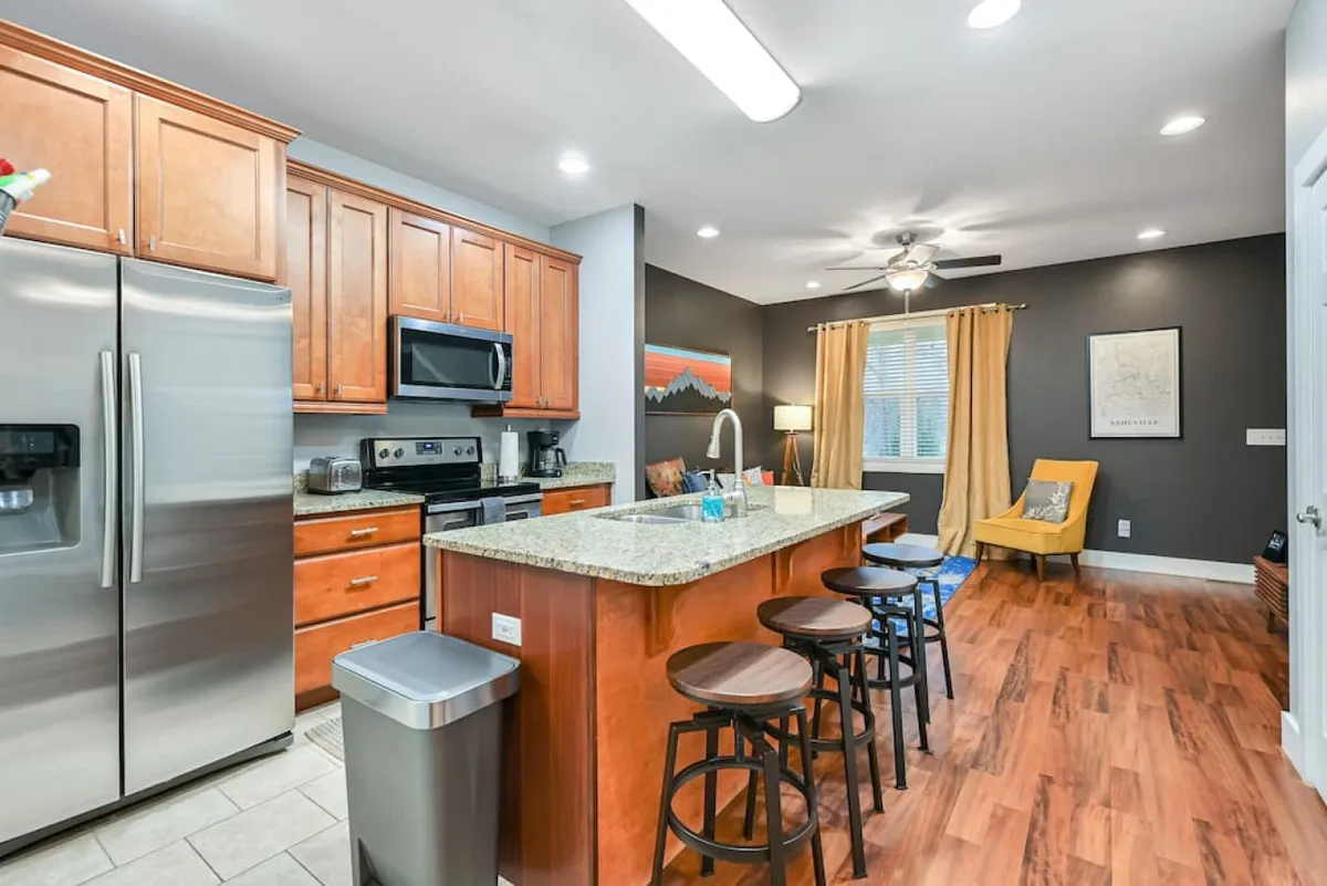 The image showcases a spacious, contemporary kitchen with a large island at its center, gleaming stainless steel appliances, and vibrant wall art that adds a pop of color. Comfortable seating invites guests to gather for meals or conversation, and the entire space exudes warmth and hospitality, perfect for group activities like cooking, dining, and socializing.