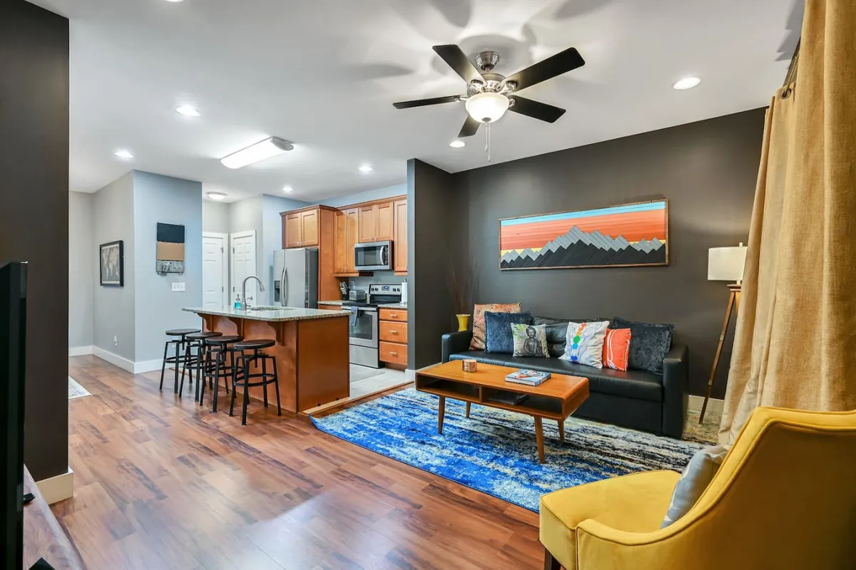 This image captures a welcoming open-concept living area, featuring a cozy sofa adorned with colorful cushions, a wooden coffee table, a bright blue area rug, a yellow accent chair, and, importantly, a view extending toward the kitchen area. The space is filled with natural light from large windows, and the arrangement encourages both relaxation and social interaction.