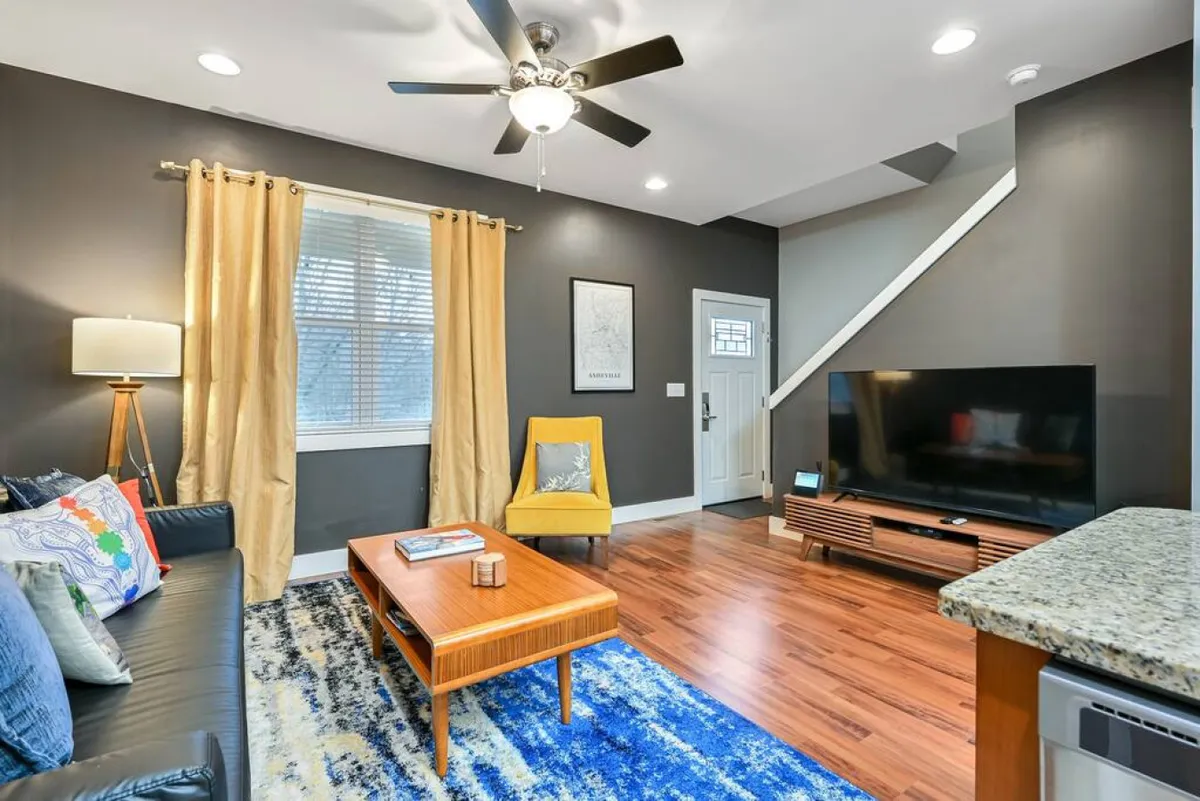 A cozy living room setting with a plush sofa, coffee table, inviting yellow chair, and a television. Soft curtains filter daylight, and the space is arranged to encourage relaxation, reading, or enjoying quiet leisure time.