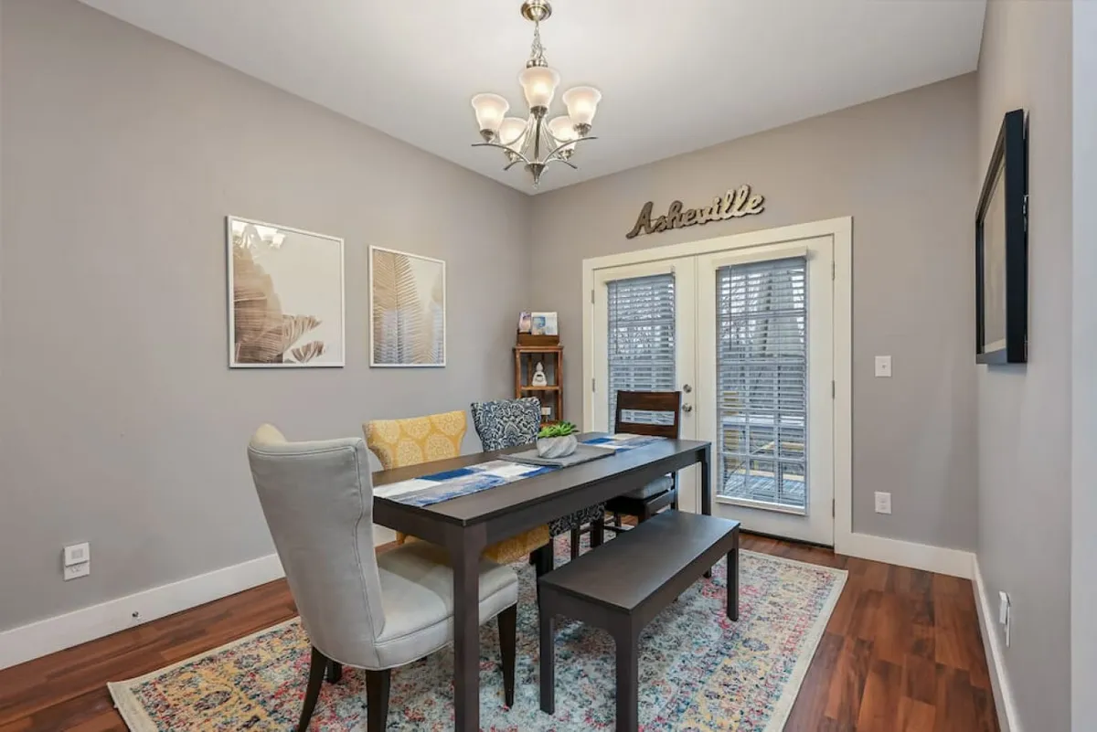 A cozy dining area with a dining table beneath a chandelier, flanked by a window with blinds and decorative artwork, including an 'Asheville' sign. The space is set up for group meals, casual gatherings, or game nights, referencing shared experiences.