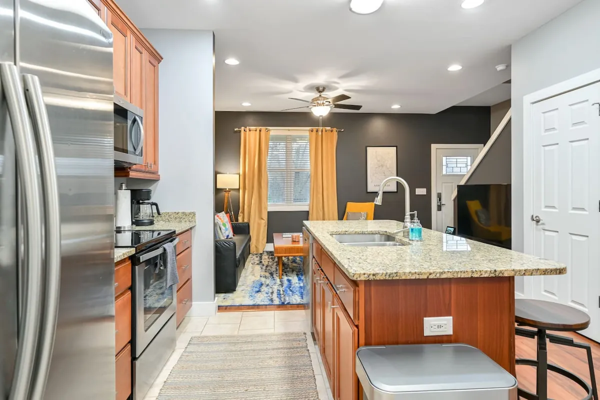 A bright, modern kitchen featuring a granite island, stainless steel appliances, colorful curtains, and a comfortable seating area. The space is well-lit and inviting, designed for both meal preparation and casual gatherings.
