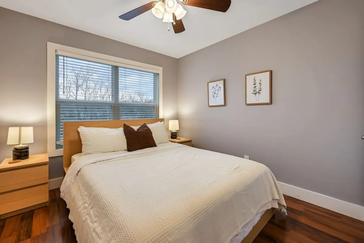 A well-appointed queen bedroom featuring a plush bed adorned with decorative cushions, flanked by nightstands with lamps, and accented by framed artwork. Soft natural light streams in, creating a peaceful and inviting atmosphere ideal for relaxation.