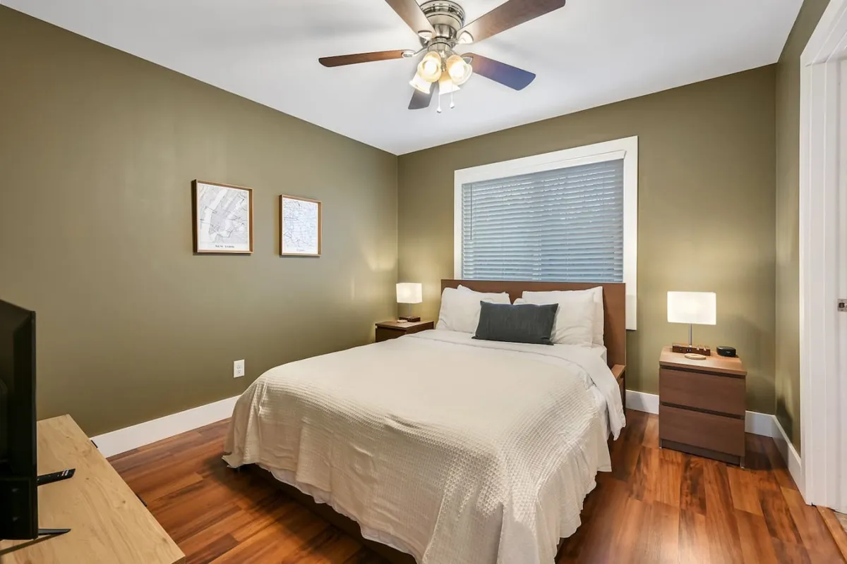 A cozy bedroom with a queen bed, nightstands, a ceiling fan, and wall artwork. The neutral color palette and soft lighting evoke a restful and calm environment, emphasizing relaxation and personal space.