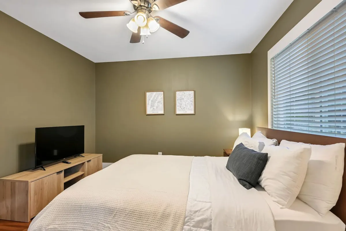 A tranquil bedroom scene featuring a bed with crisp white linens, elegant wooden furniture, a television on a stand, and thoughtfully chosen framed artwork on the wall. Soft natural light filters in, highlighting the calm ambiance of the space.