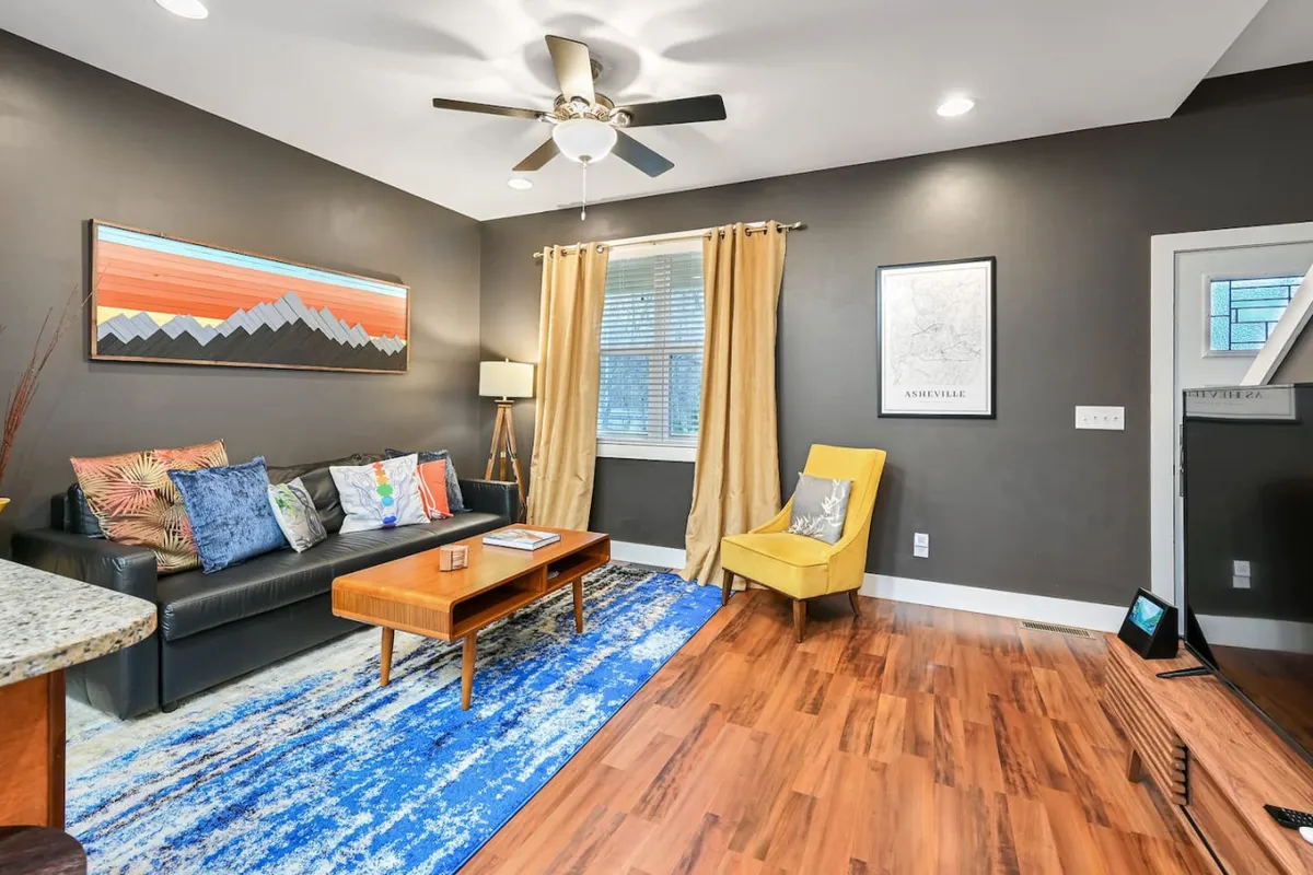 A cozy living area with a stylish sofa, coffee table, vibrant artwork, a window letting in natural light, and a cheerful yellow chair. The space is arranged for relaxation, socializing, and enjoying quiet moments.