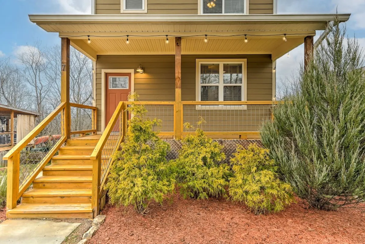 This image captures the exterior of the home, featuring a welcoming wooden staircase, the front door, and lush green shrubs. The scene conveys a sense of arrival and hints at the inviting, social atmosphere within.