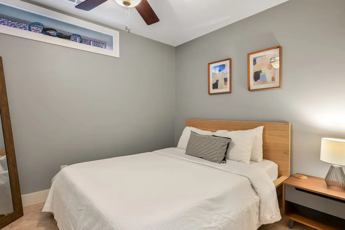 A calm and inviting bedroom with white linens, two pieces of framed artwork above the bed, and a nightstand with a glowing lamp. Gentle sunlight streams into the room, creating a soft, peaceful mood ideal for relaxation.