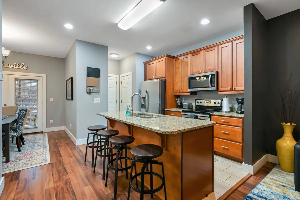 This image features a modern kitchen with granite countertops, stainless steel appliances, warm wooden cabinetry, and bar stools at the counter. The open layout invites group meal preparation and casual dining, with space for guests to gather and converse.